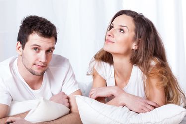Ein junges Paar liegt auf die Ellenbogen gestützt im Bett und sieht fragend in die Kamera